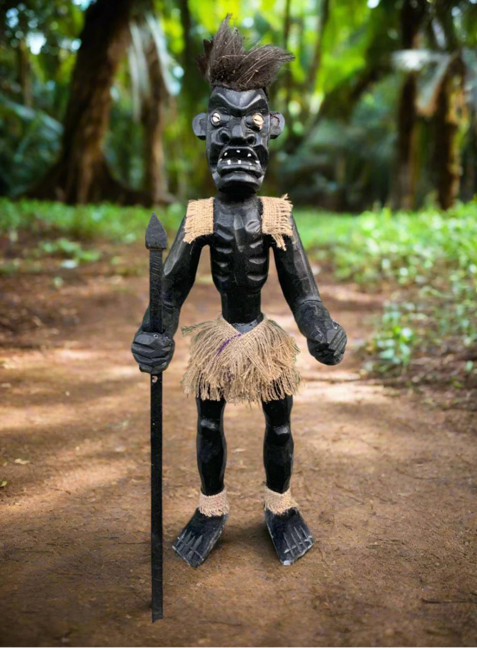 Carved black wooden tribal figure with burlap skirt and shoulder wrap, holding a spear in a forest setting