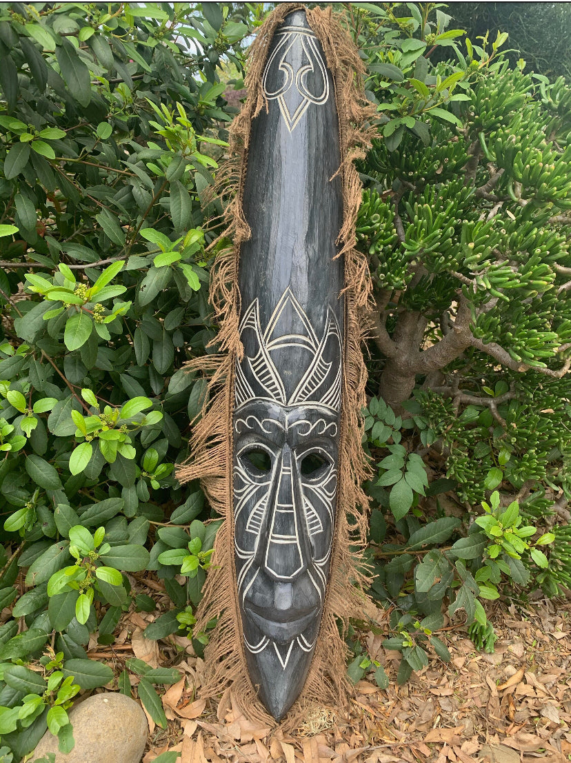 Elongated wooden tribal mask with white geometric patterns and a jute fringe among green plants