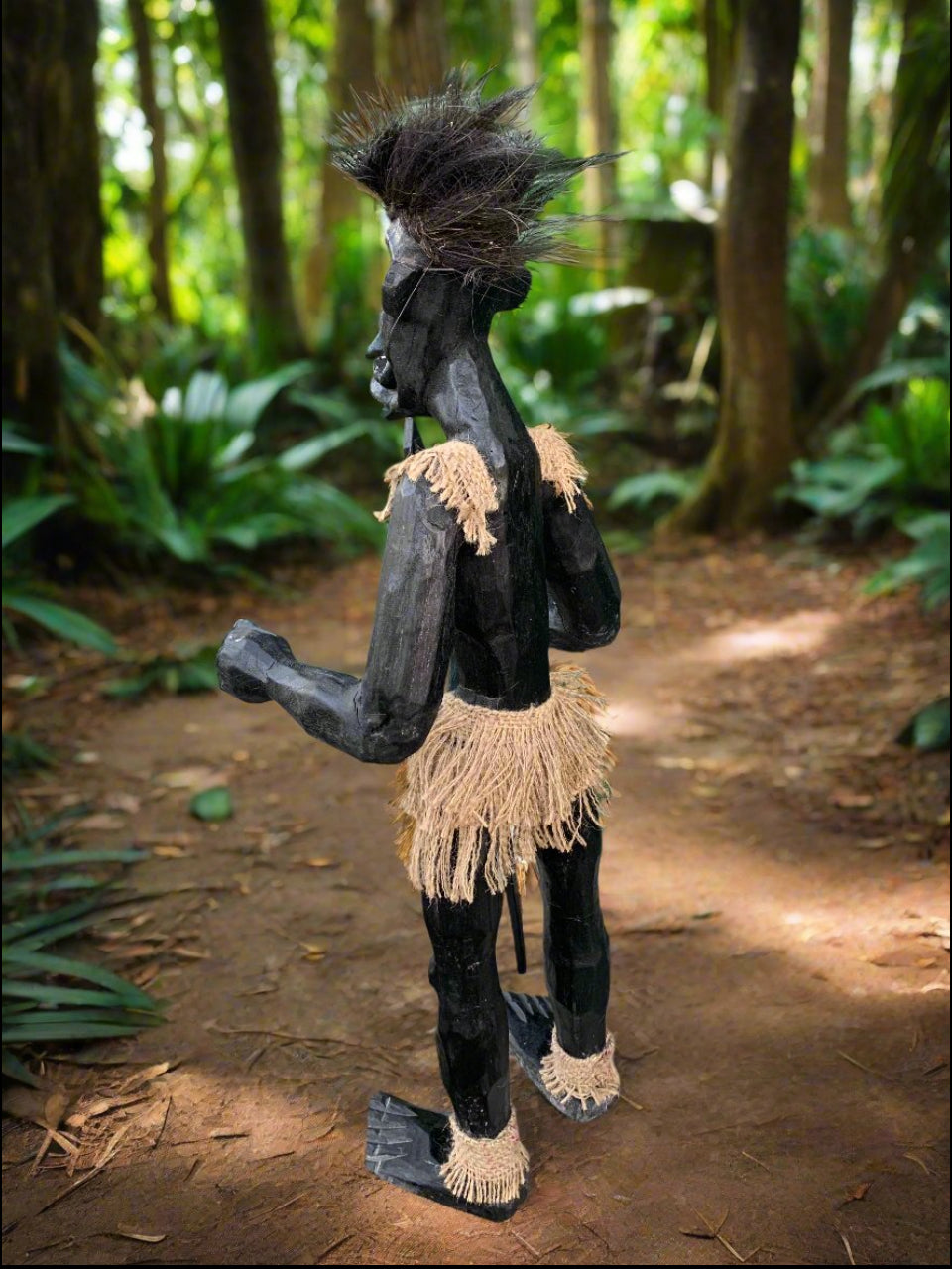 Black wooden tribal figurine with straw fringe and spiky hair in a forest path