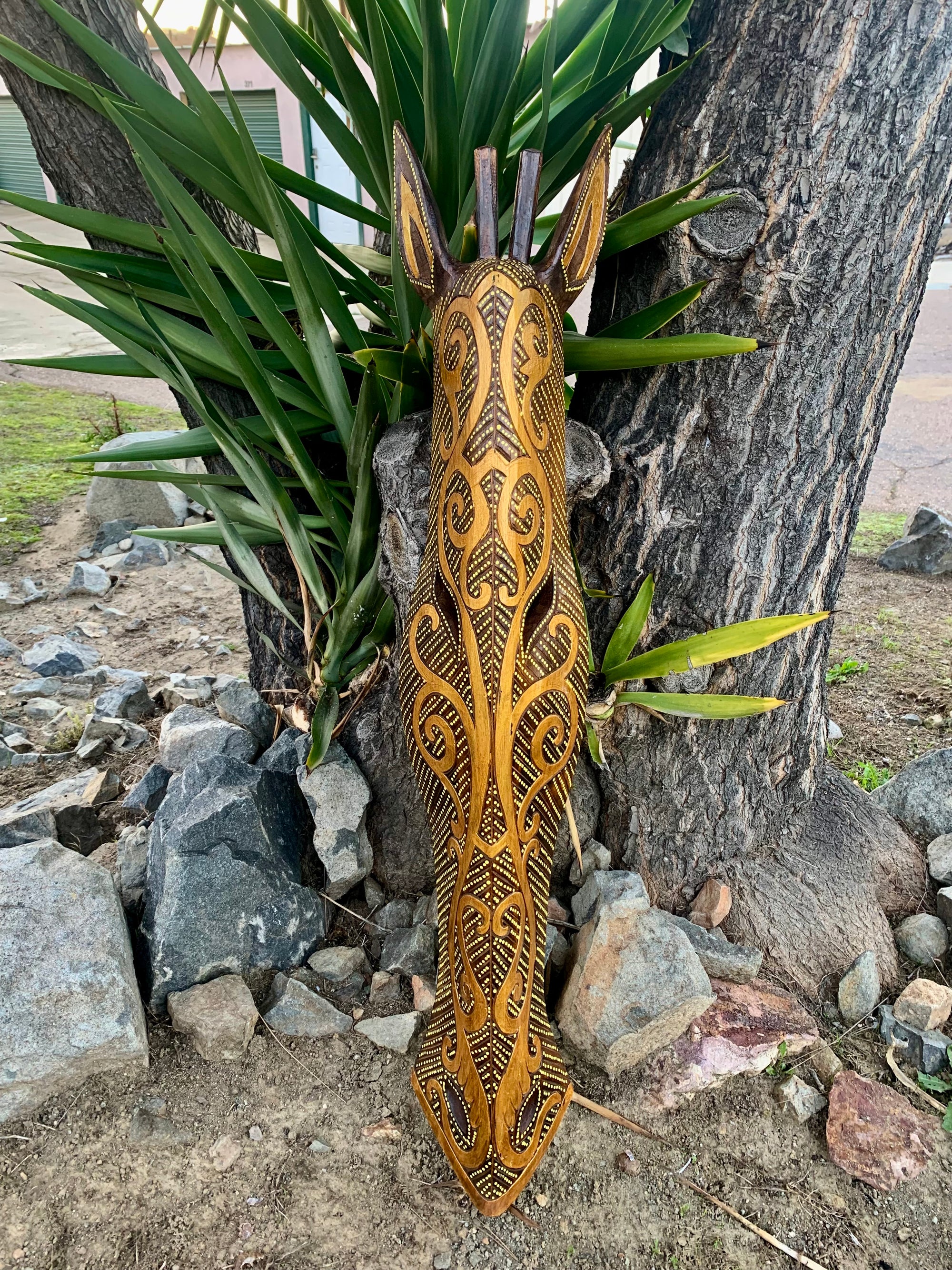 Tribal-patterned wooden giraffe sculpture leaning against a tree outdoors