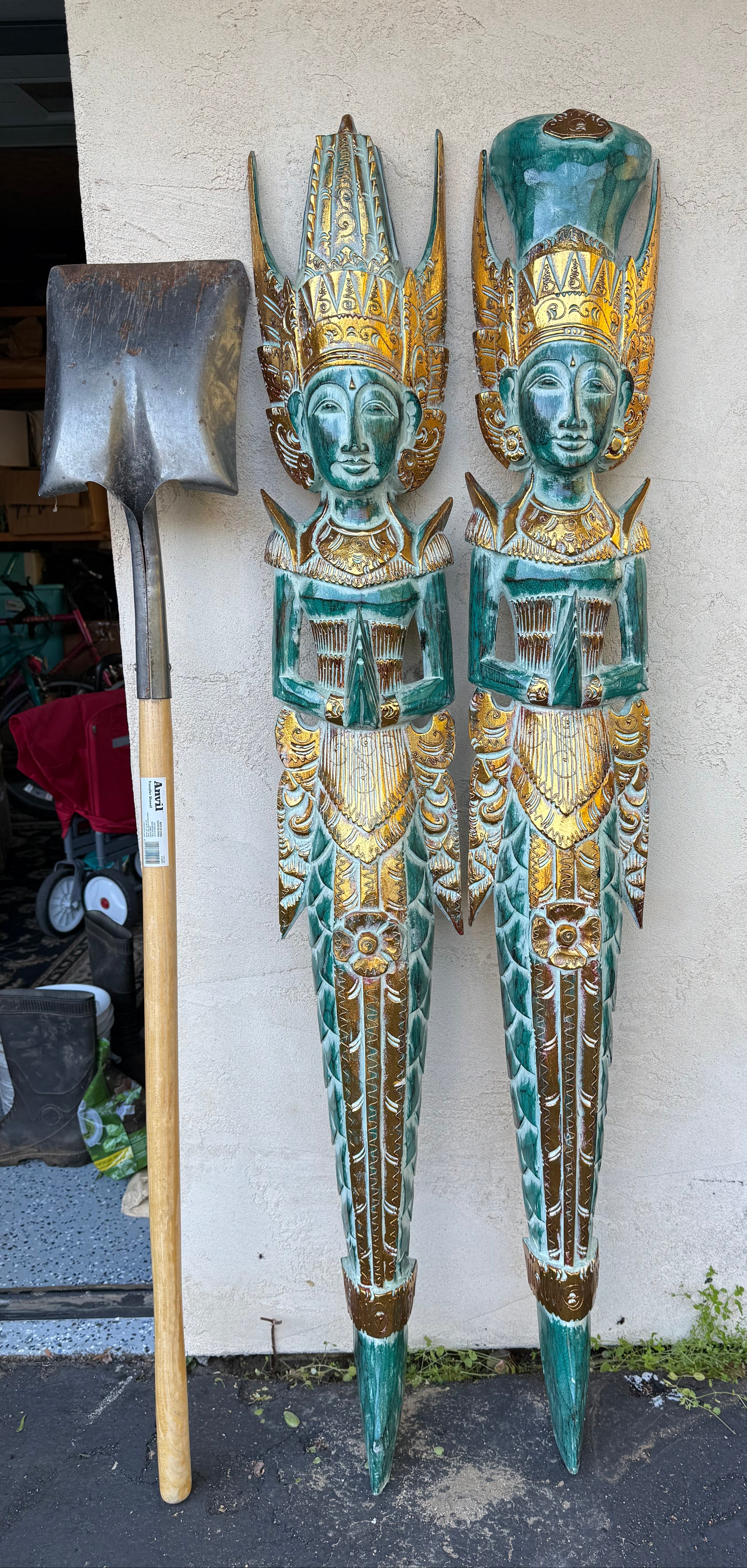 Two turquoise and gold carved wooden statues with praying hands beside a shovel against a wall