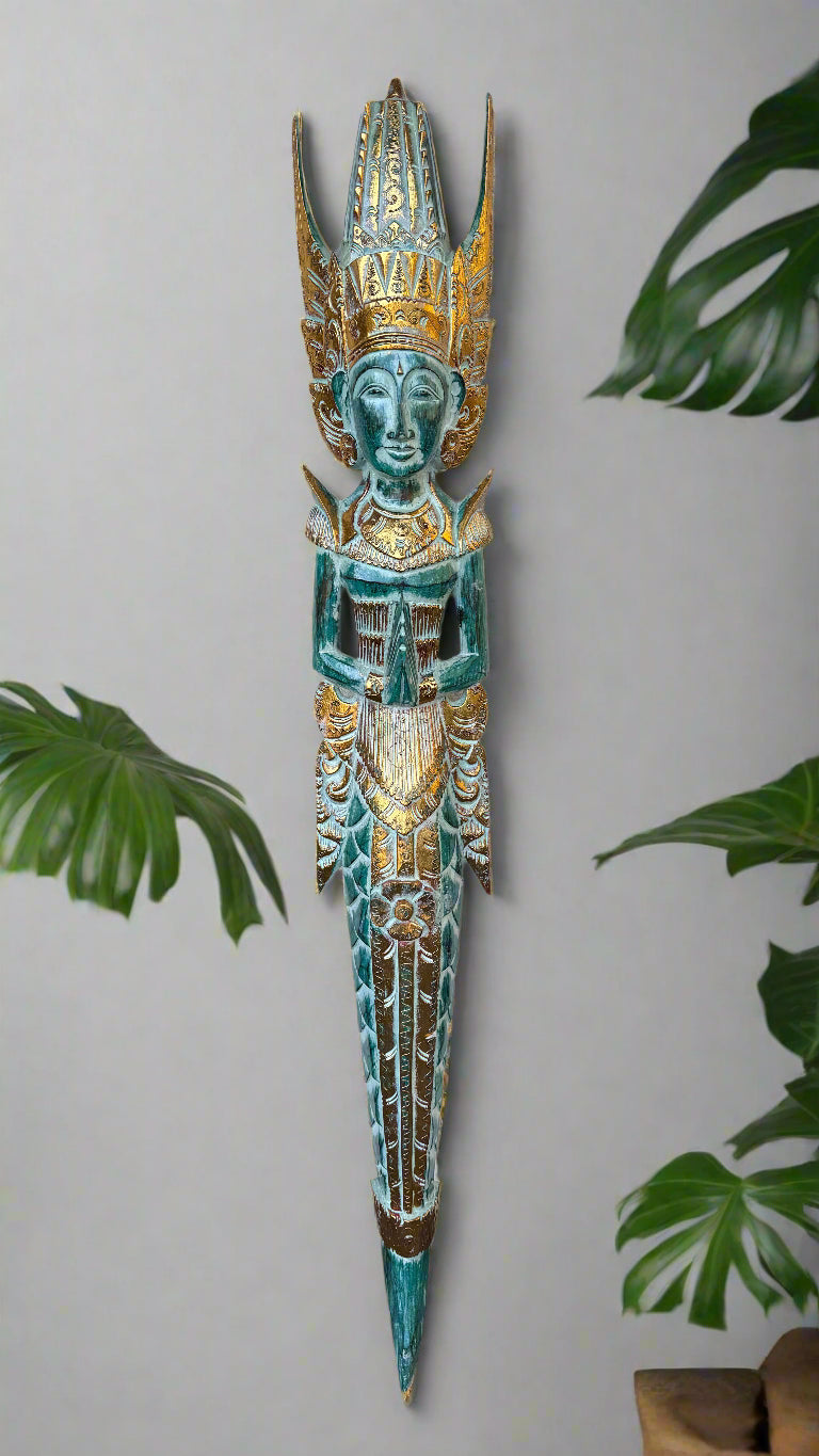 turquoise-and-gold ornate carved statue with crown and folded hands against a neutral wall