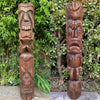 Two carved wooden tiki totems with expressive faces standing outdoors among green shrubs.