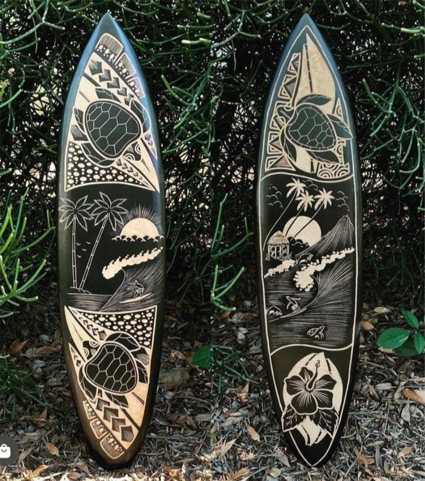 Two carved surfboards with tropical island scenes resting against green shrubbery.