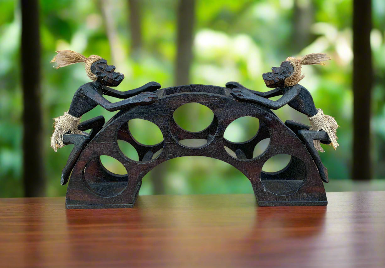 Two carved black wooden tiki figures pushing a circular arched sculpture with cut-out holes.