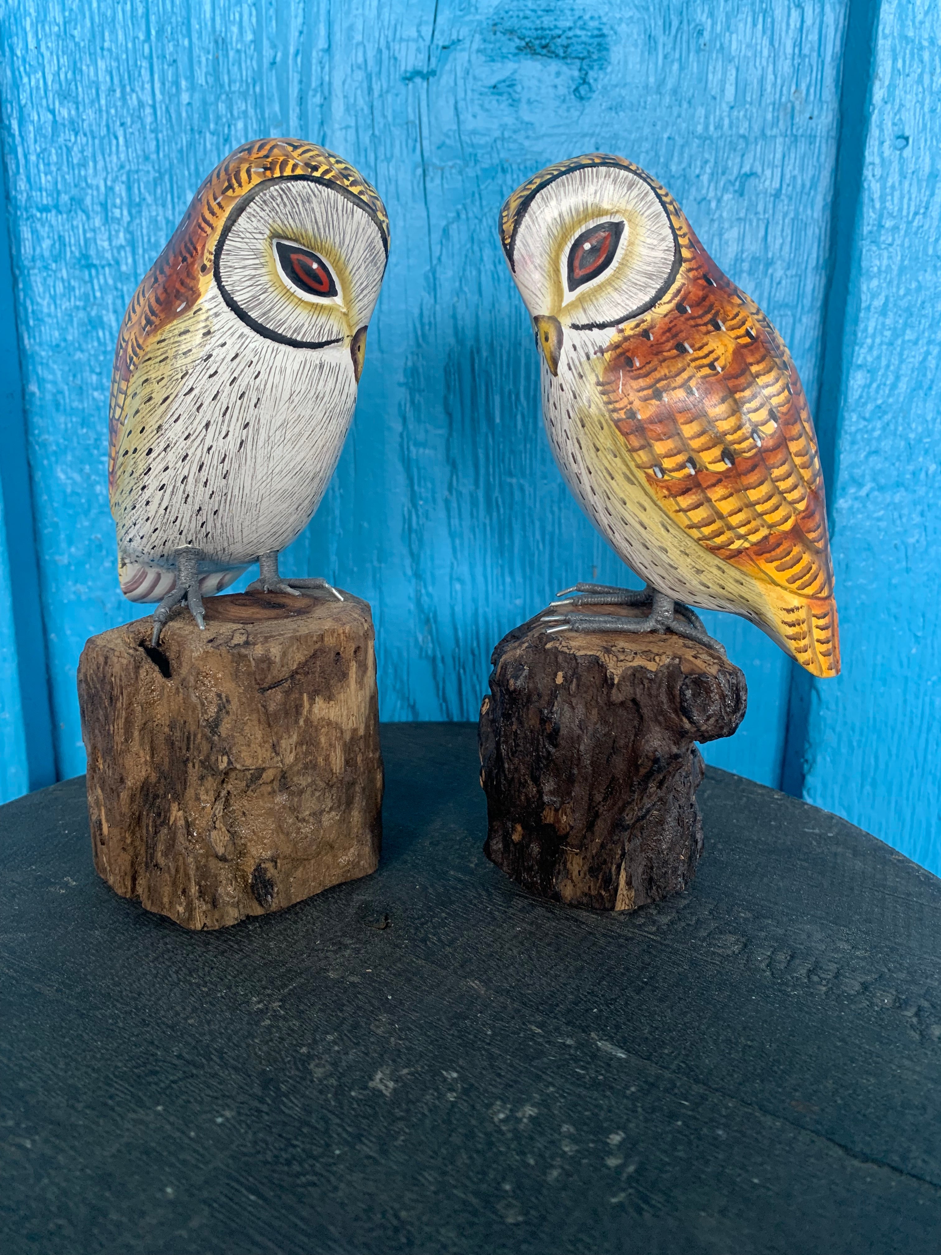 Two carved wooden owl figurines perched on small tree stump bases against a bright blue wooden background.