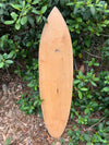 Unfinished wooden longboard leaning against a green hedge.