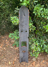 Tall weathered wooden garden sculpture with scalloped edges and circular cutouts leaning against green shrubs.