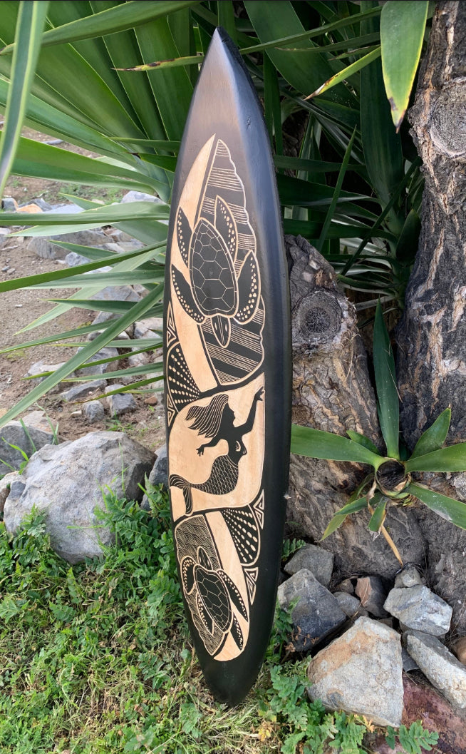 Woodburned longboard with mermaid and lotus motifs against rocks and greenery