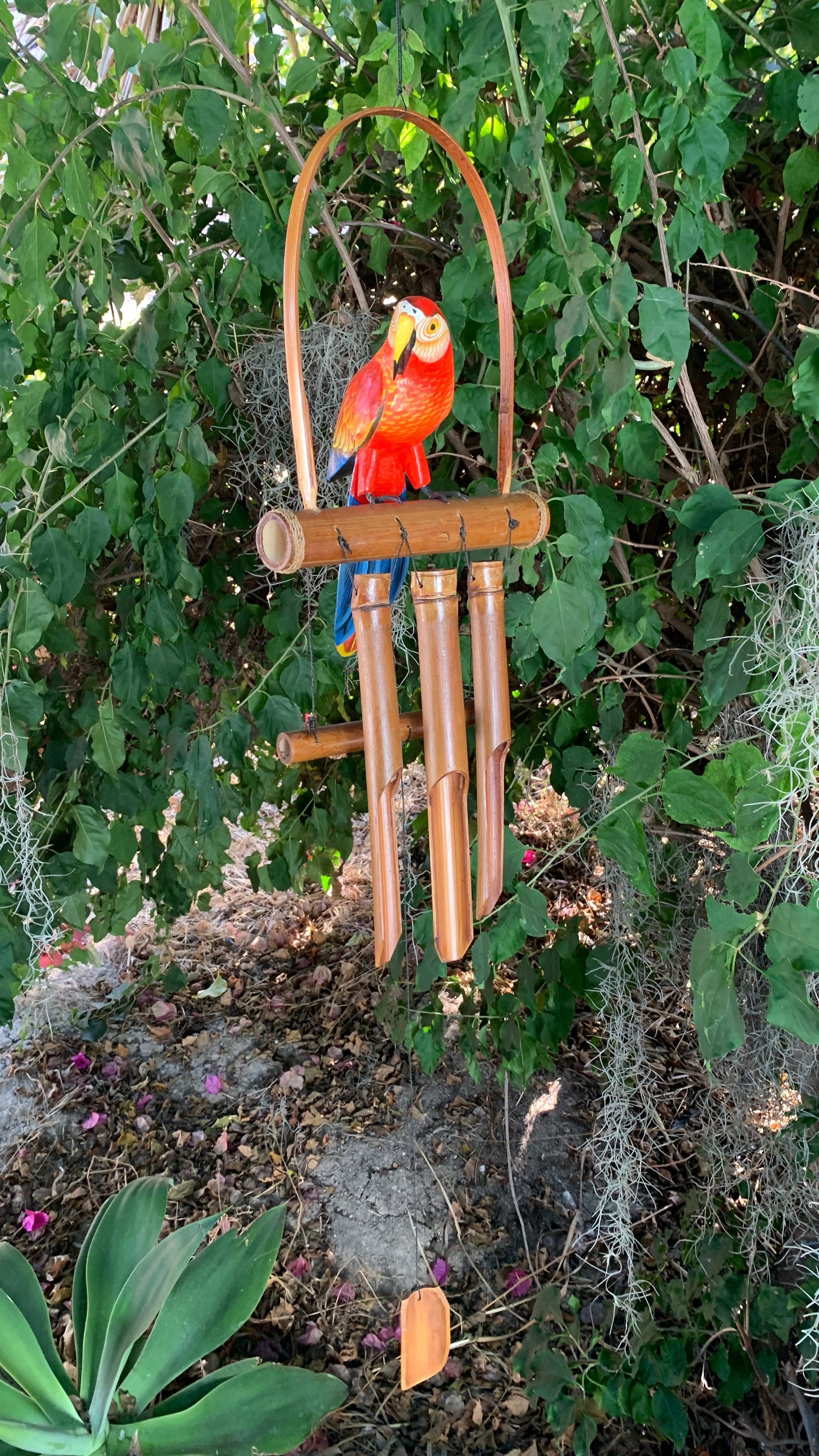 Bright red-orange parrot perched on a wooden bamboo wind chime among green leaves