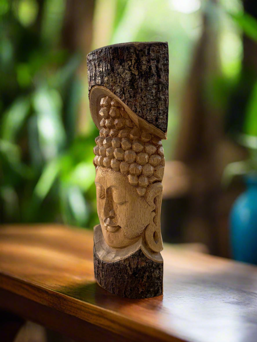 Wooden Buddha head carving with textured curls on a tree trunk, placed on a wooden table with a blurred green background.