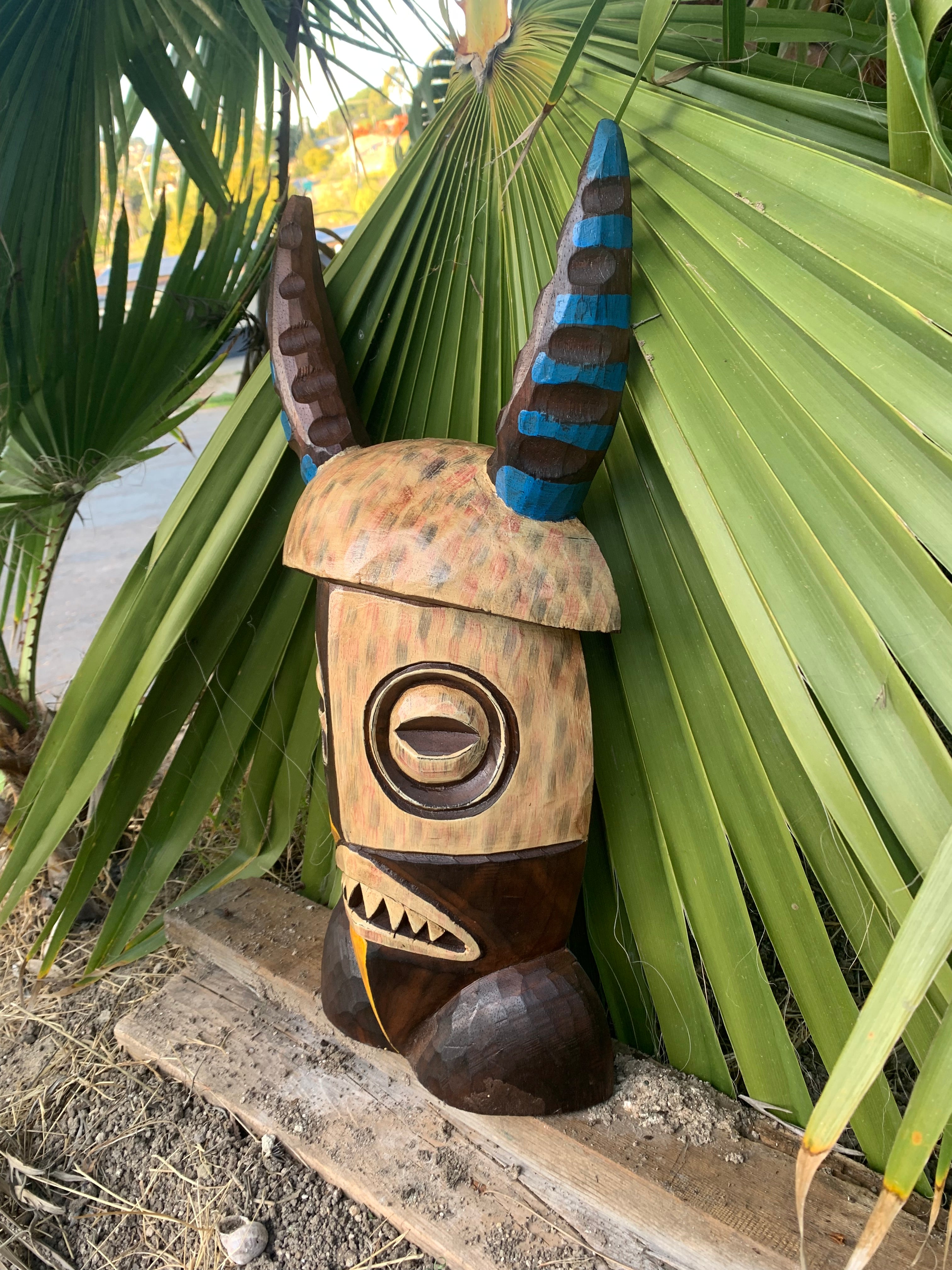 Wooden carved mask with horned headdress and blue stripes among palm leaves