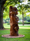 Wooden carved robed statue standing on a circular base in a sunny park.