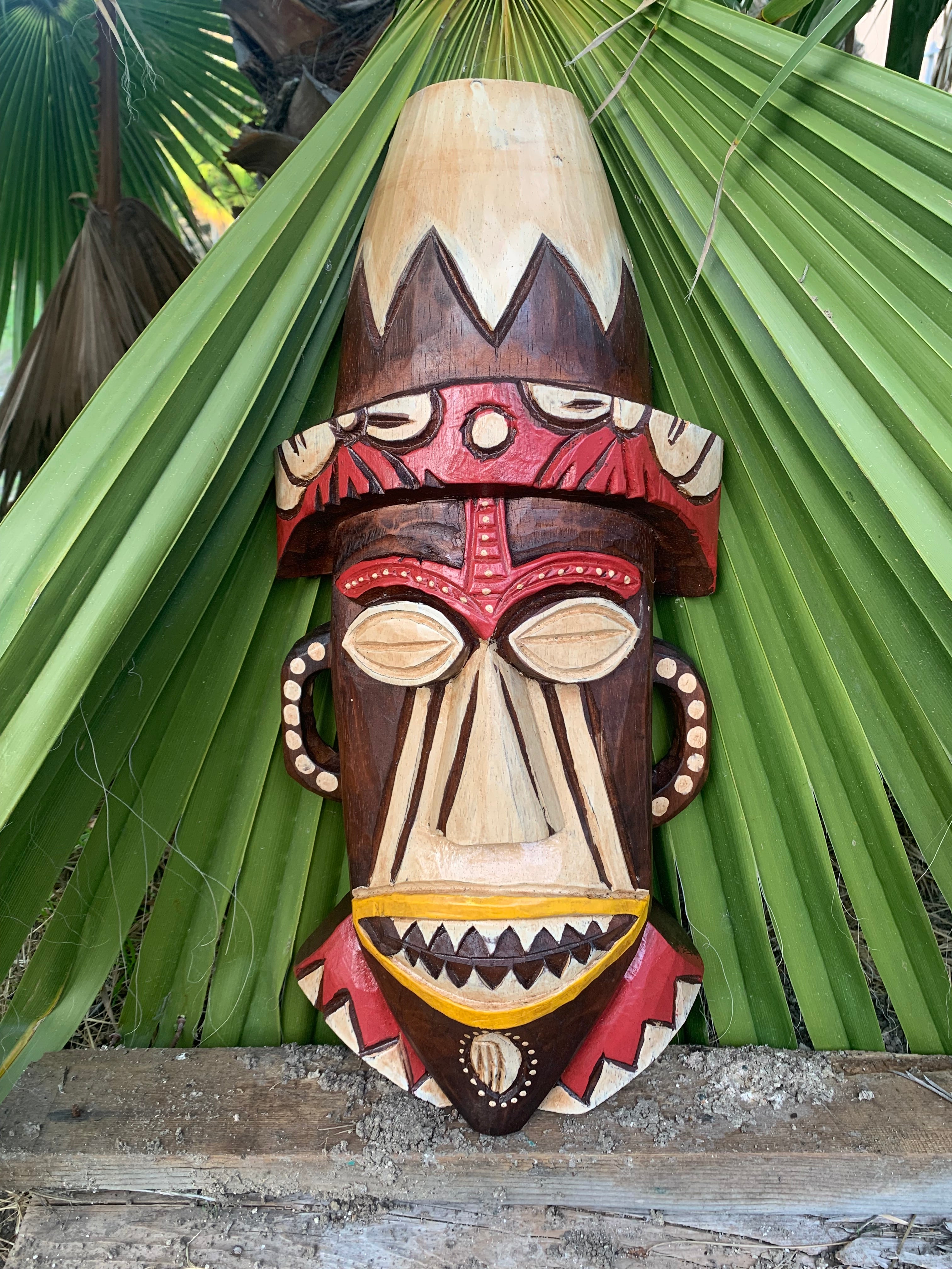 Wooden carved tiki mask with headdress, brown and cream colors, leaning against green palm leaves.