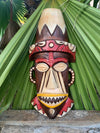 Wooden carved tiki mask with headdress, brown and cream colors, leaning against green palm leaves.