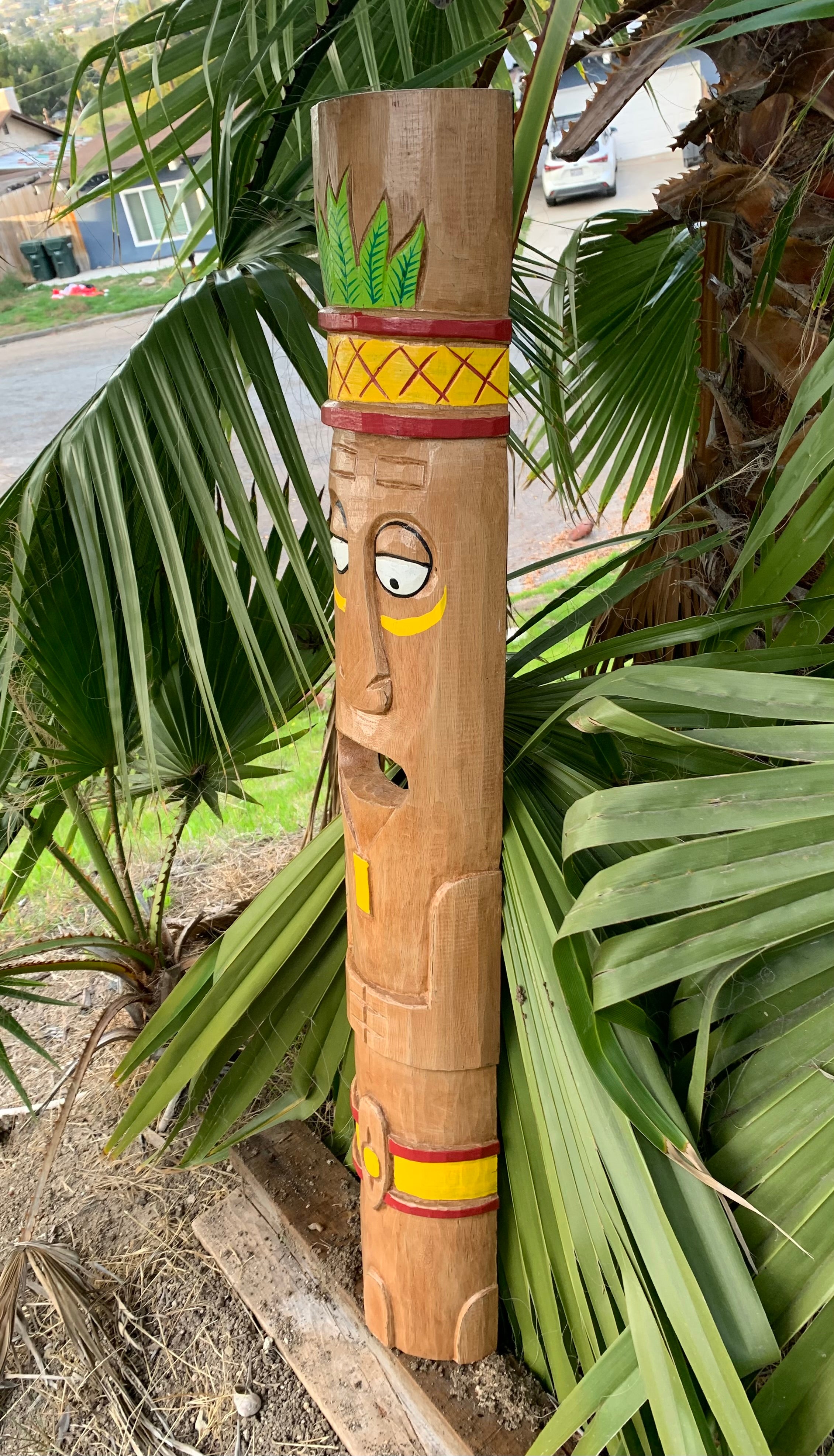 Wooden carved totem pole with a face and colorful red and yellow bands beside palm leaves