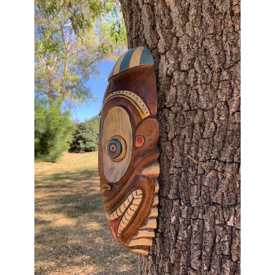 Wooden carved tribal mask mounted on a tree in a park.