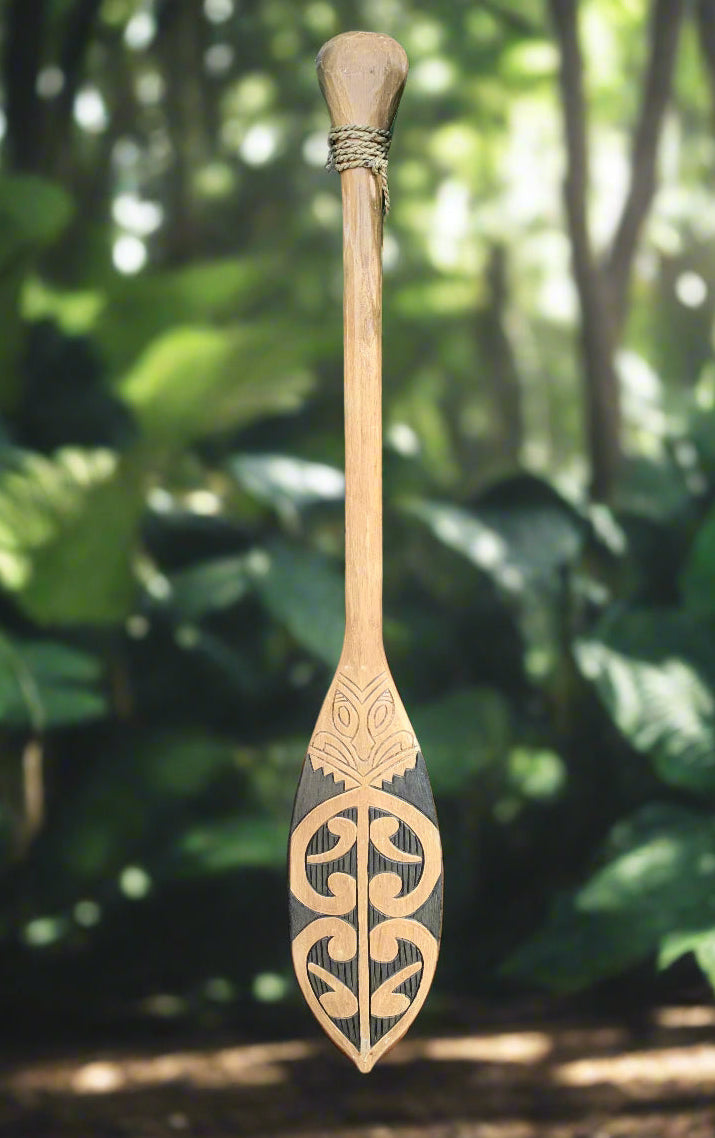 Carved wooden paddle with Maori koru patterns and rope-wrapped handle in a forest setting