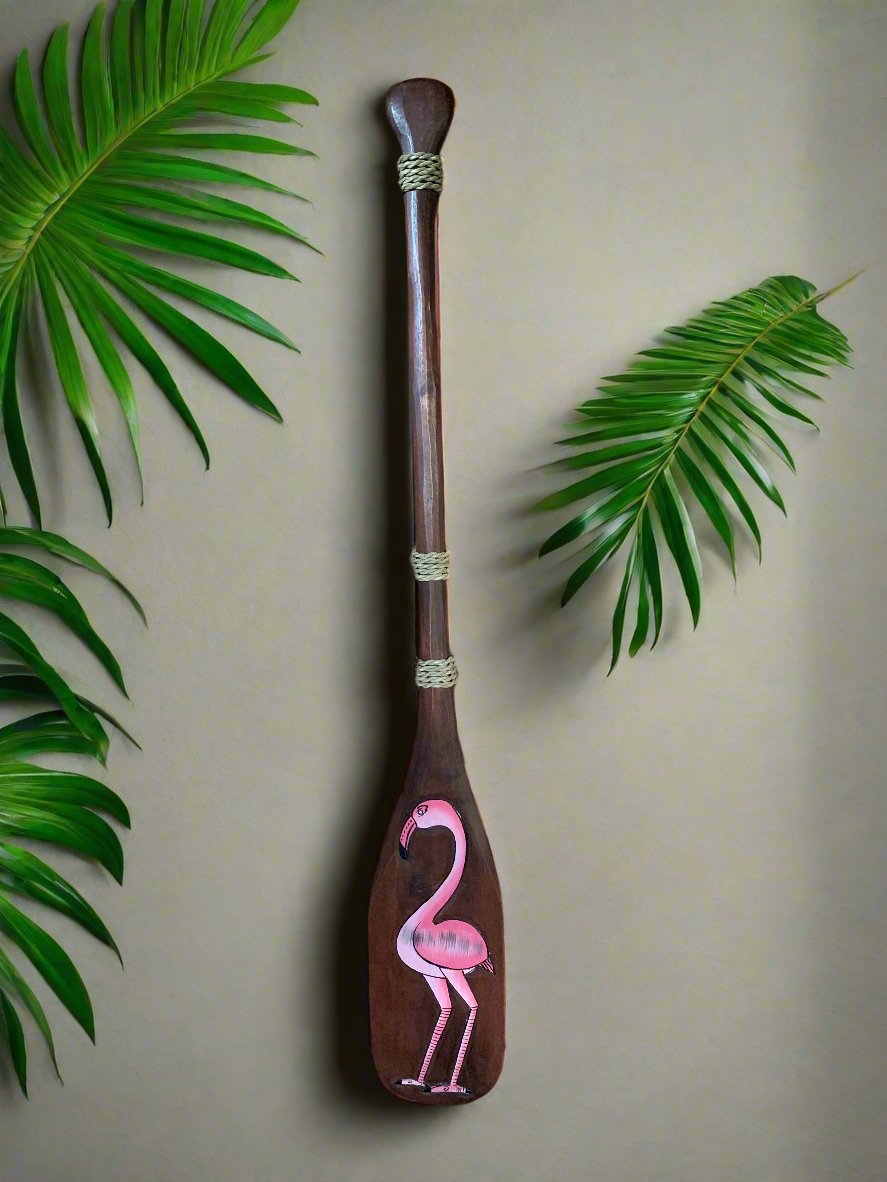 Brown wooden paddle with a pink flamingo painting, surrounded by tropical green leaves.