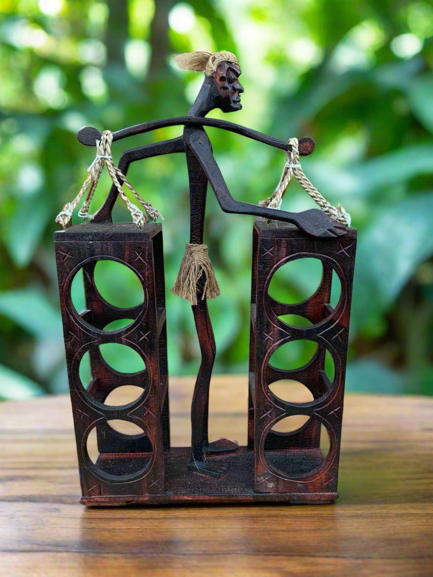 wooden sculpture of a slender figure balancing two tall towers with circular cutouts outdoors