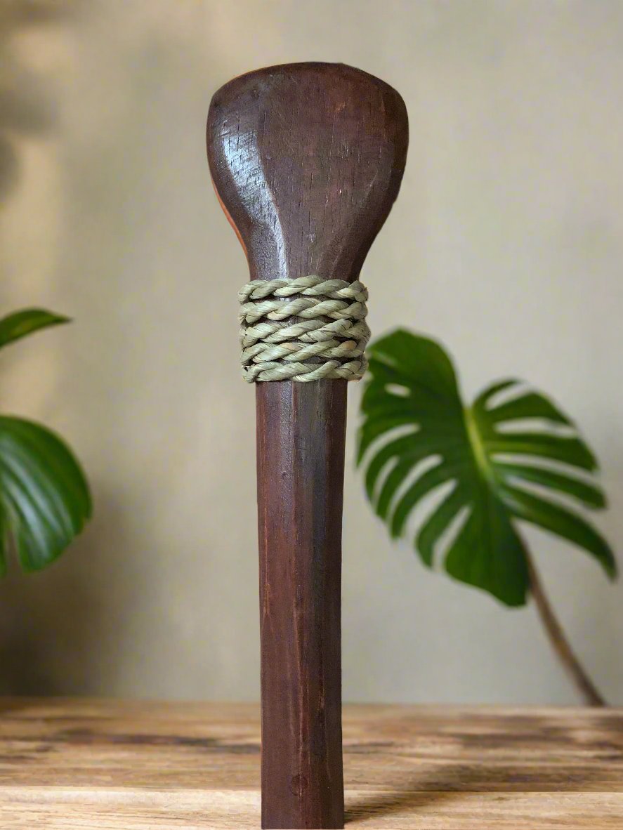 Dark wooden staff with braided rope bound near the top, on a wooden surface with green plants in the background.