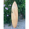 Light wooden surfboard leaning upright beside a green hedge with pink morning glory flowers
