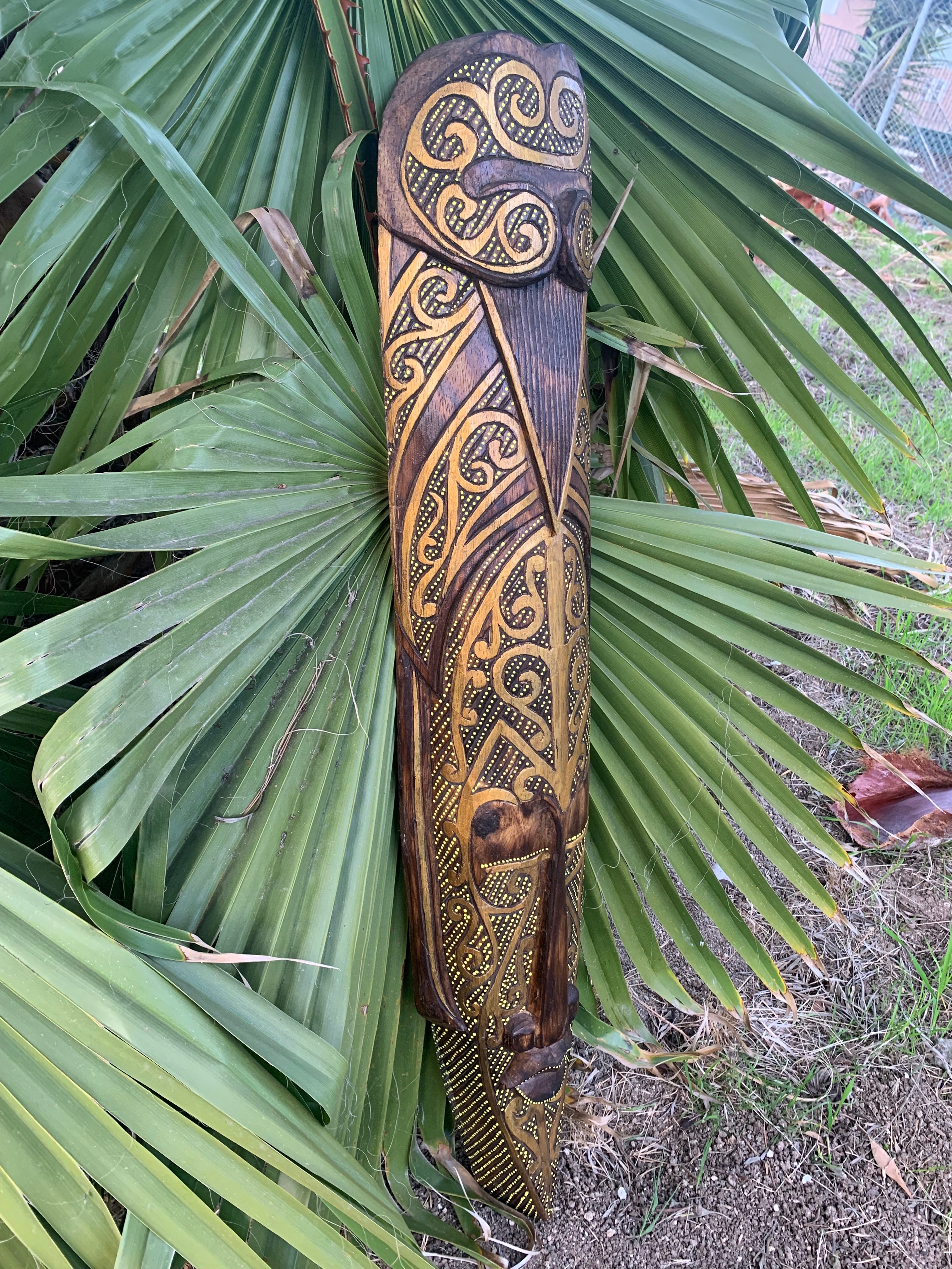 Intricately carved tiki with gold ornamentation and swirl patterns on dark wood beside palm fronds.