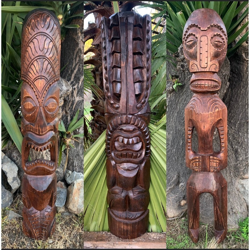 Three carved wooden tiki statues in a garden setting with plants and rocks