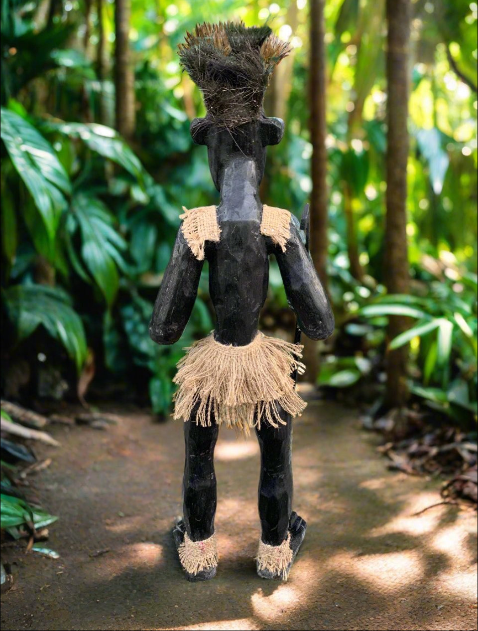 Back view of a black wooden tiki figure wearing a straw skirt in a tropical rainforest.