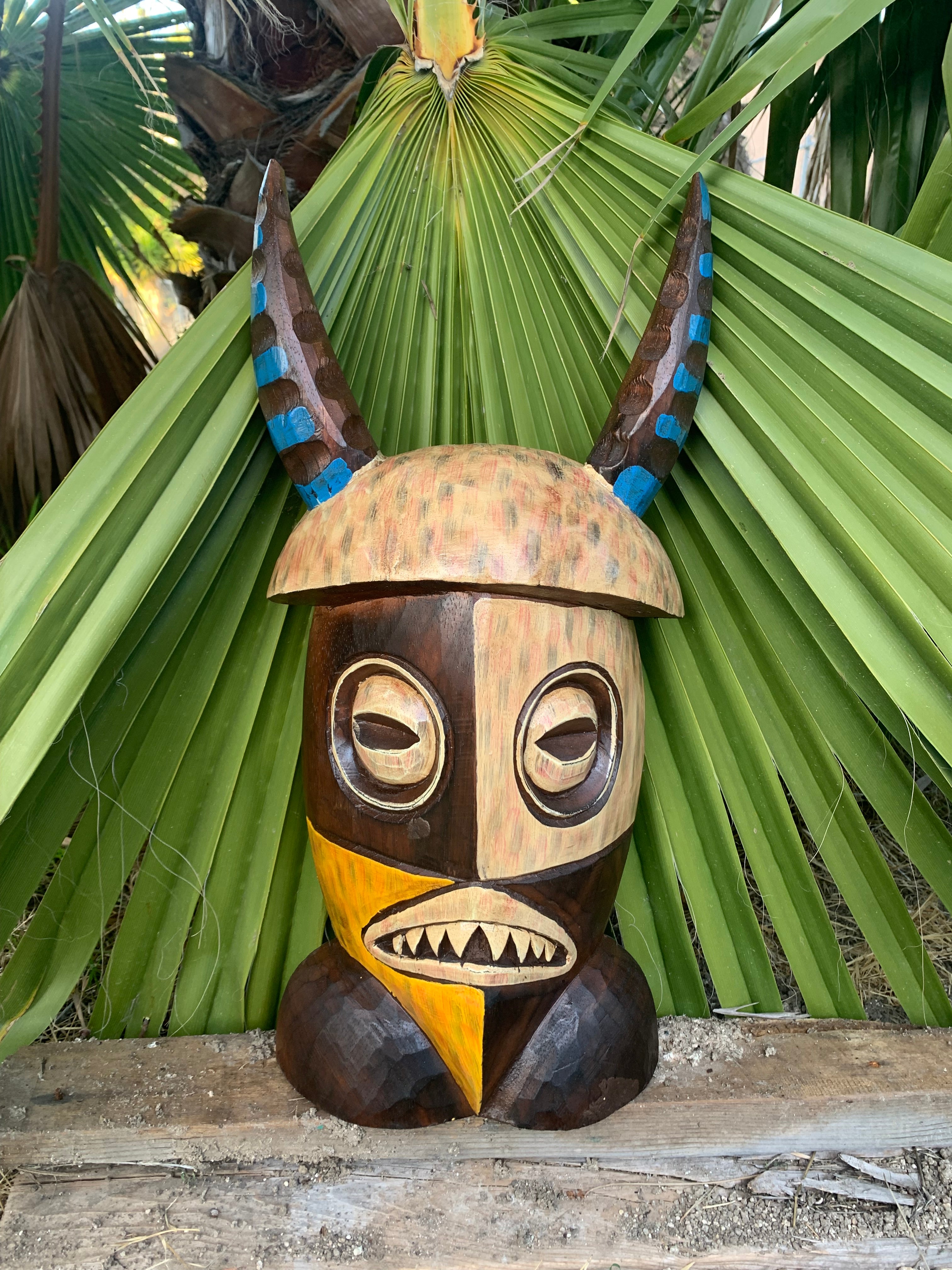 Carved wooden tiki mask with blue-accented horns, outdoors among palm leaves.