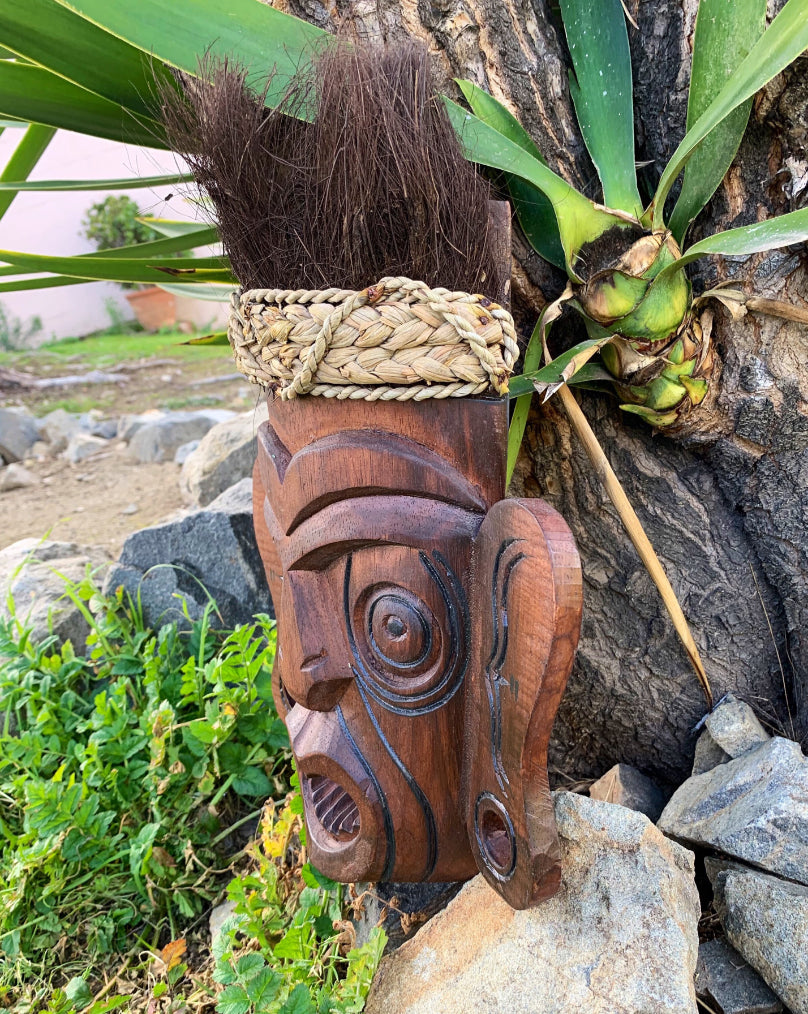 Wooden tiki mask with rope-wrapped top mounted on a tree trunk