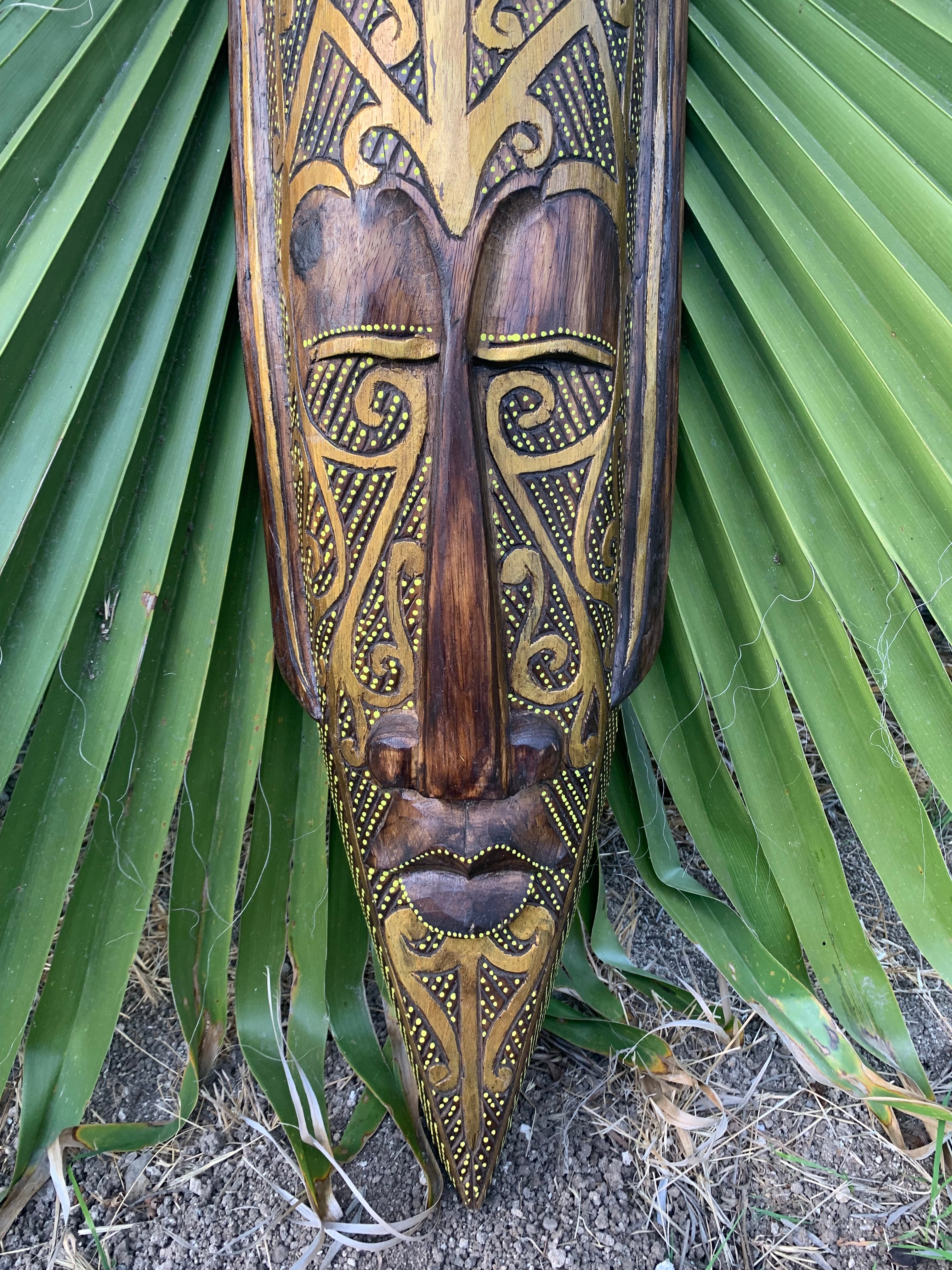 Carved wooden tiki mask with gold dotted patterns on green plant leaves backdrop