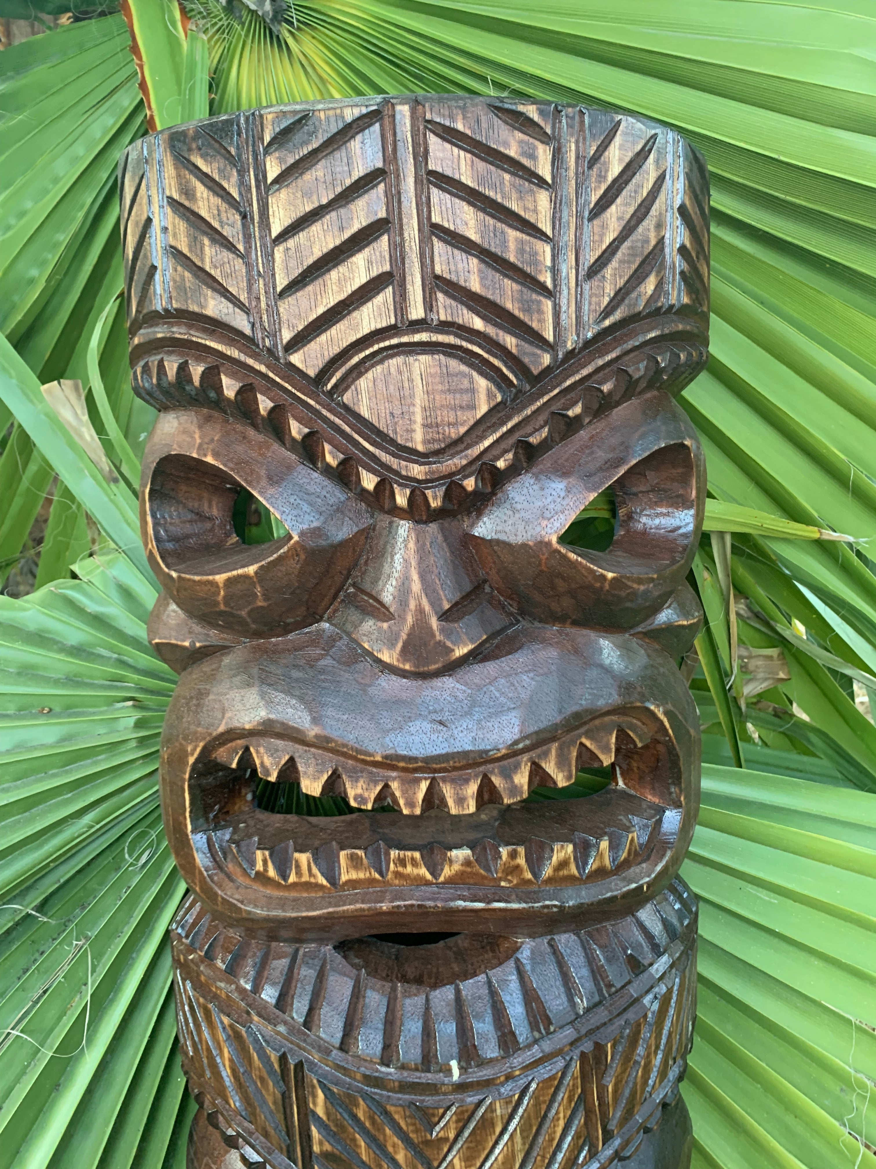Carved wooden tiki statue with geometric patterns and prominent teeth, set among tropical palm leaves.