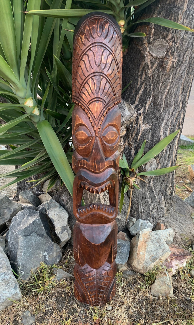 Carved wooden tiki statue with geometric patterns and open mouth beside a tree trunk