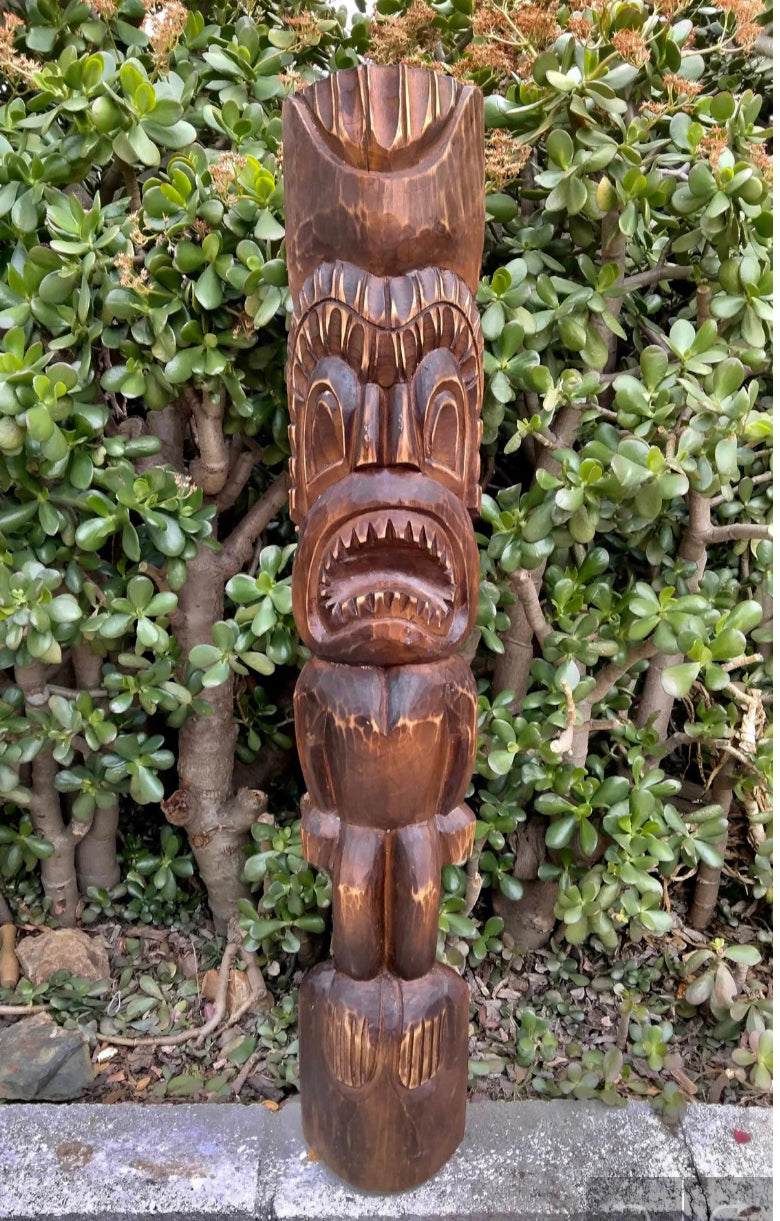 Carved wooden tiki statue with an open mouth, set against green shrubbery.
