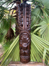 Wooden tiki totem sculpture carved with faces standing among palm leaves