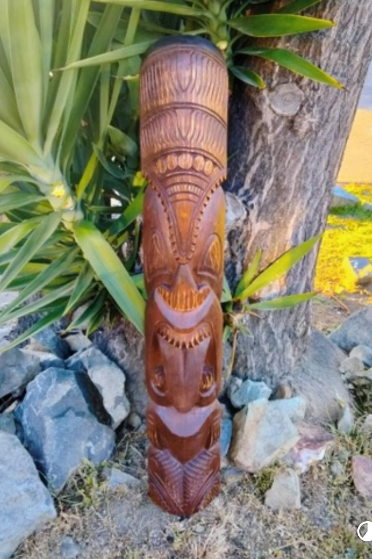 Tall wooden tiki totem pole carved with multiple faces outdoors beside rocks and greenery