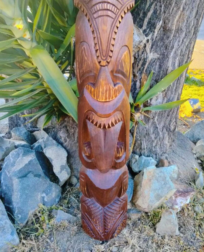 Wooden tiki-style totem carving with a smiling face outdoors in a garden setting