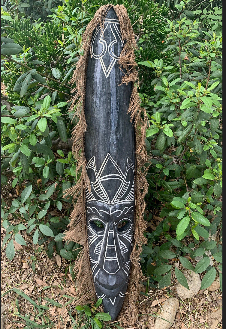 Dark wooden tribal mask with white geometric carvings and brown fringe, outdoors among green shrubs