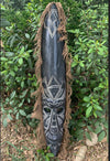 Dark wooden tribal mask with white geometric carvings and brown fringe, outdoors among green shrubs