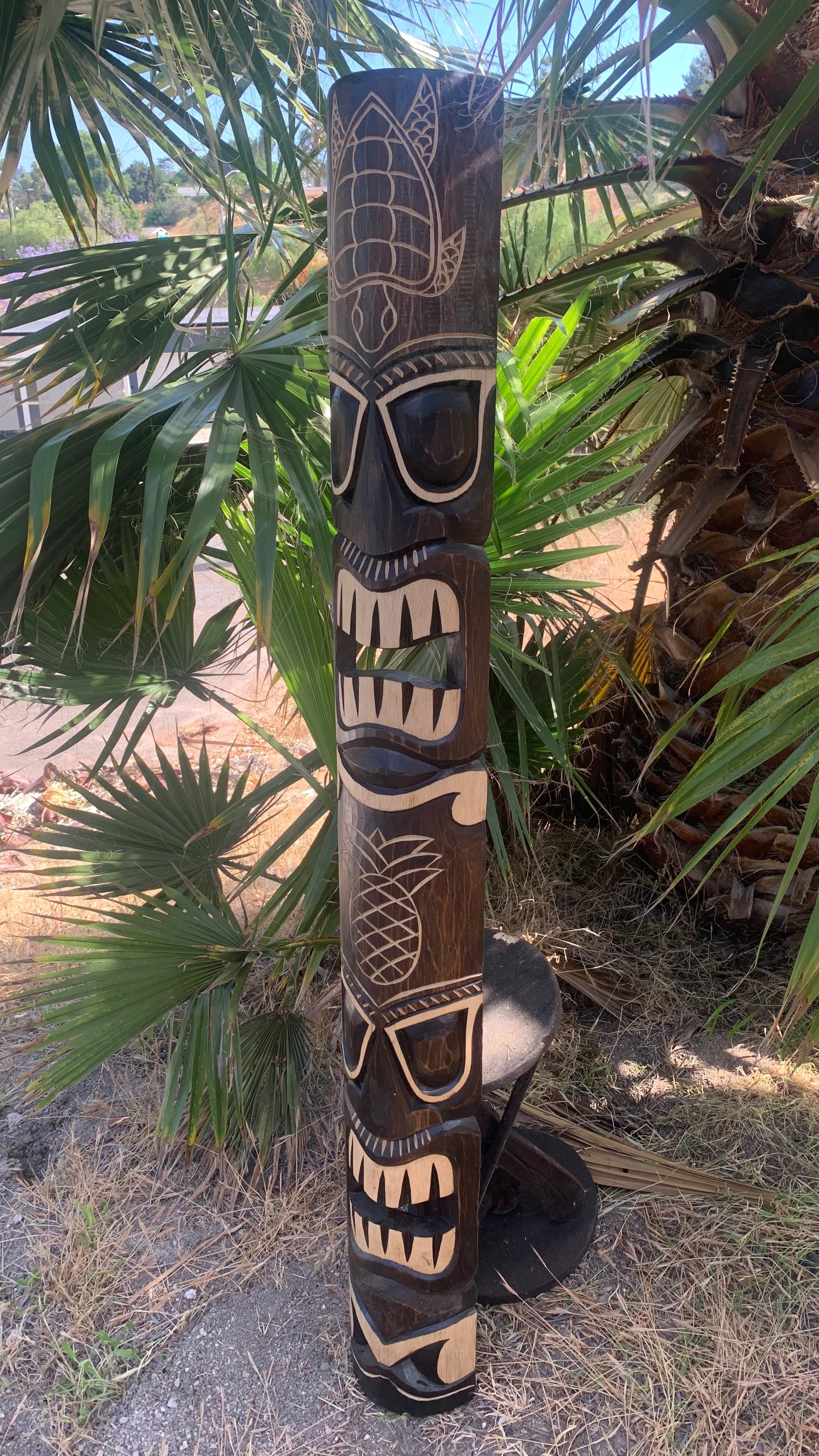 Carved wooden totem pole with black and white geometric designs outdoors among palm fronds.