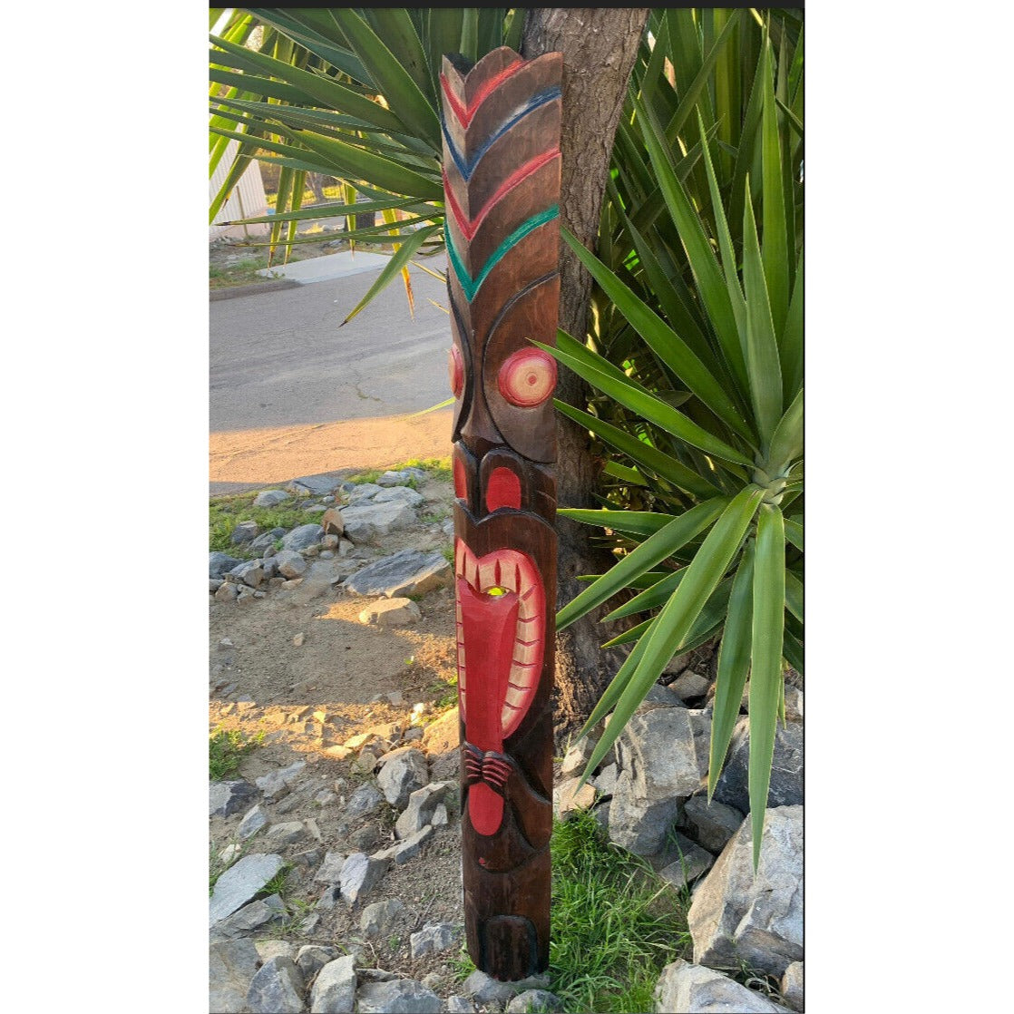 Carved wooden totem pole with red and teal designs beside a tree and green plants.