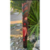Carved wooden totem pole with red and teal designs beside a tree and green plants.