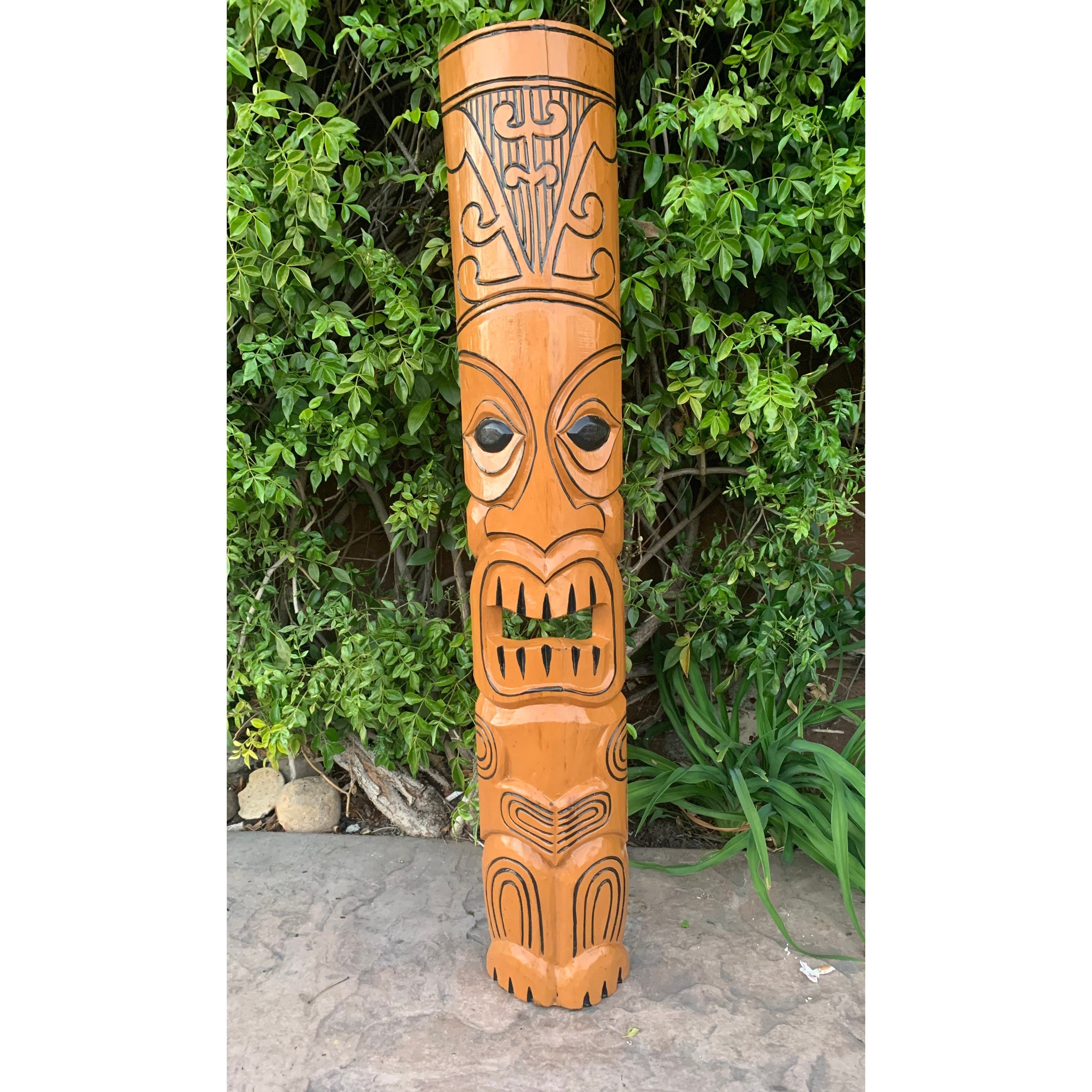 Orange wooden totem pole with a carved face and tribal patterns beside green foliage