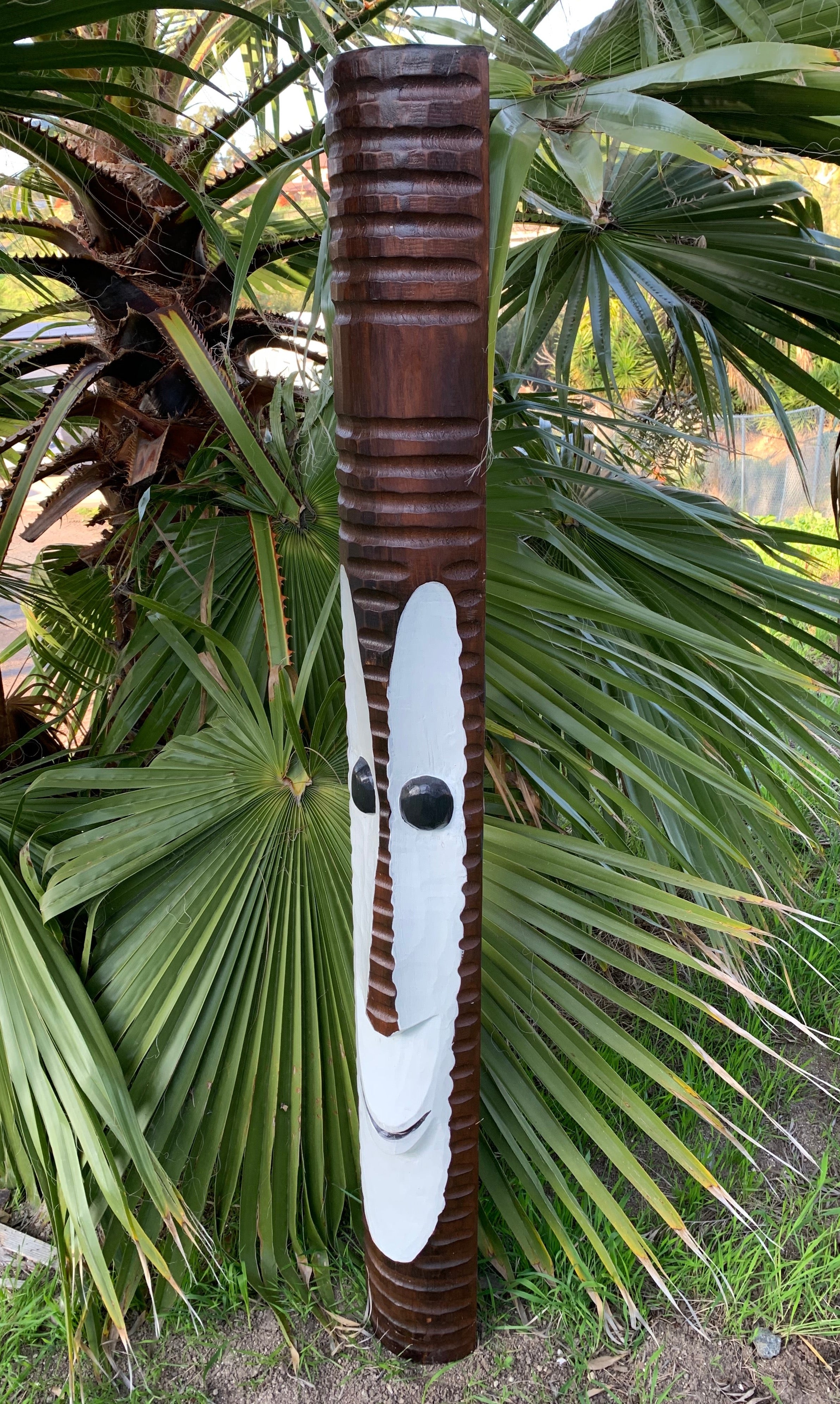 Carved wooden totem pole with a white face and black eyes in a garden among palm plants