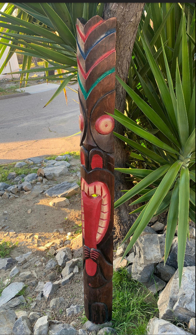 Carved wooden totem pole with red teal and blue feather patterns beside a tree