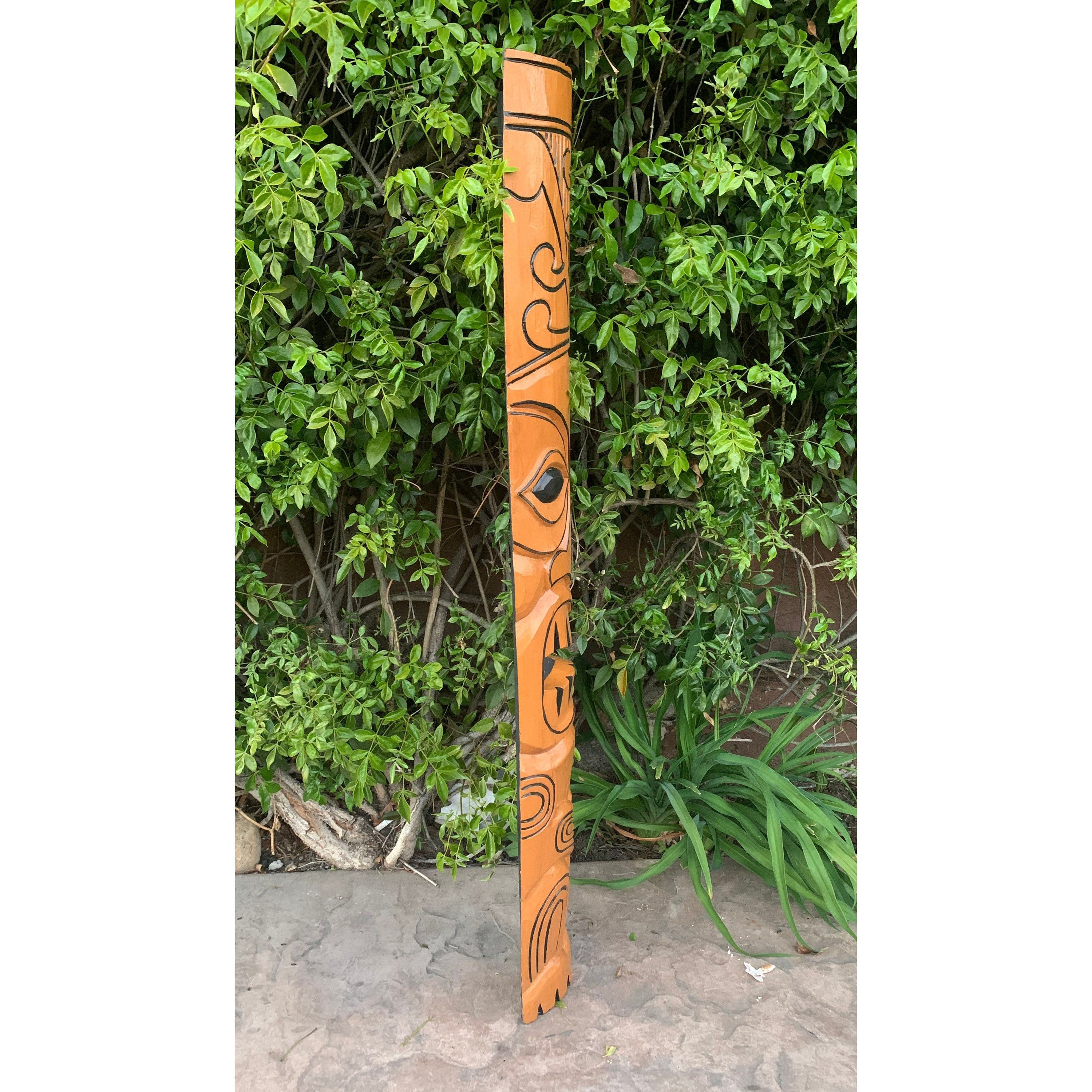 Orange wooden totem pole with black spiral designs standing near green bushes in a garden