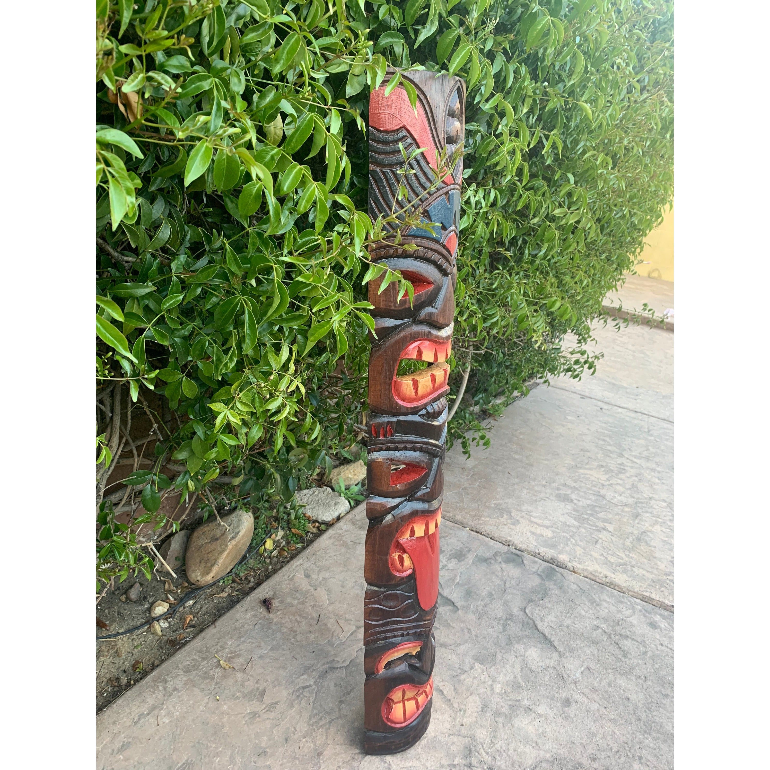 Outdoor wooden totem pole with carved faces leaning beside a green hedge along a sidewalk.