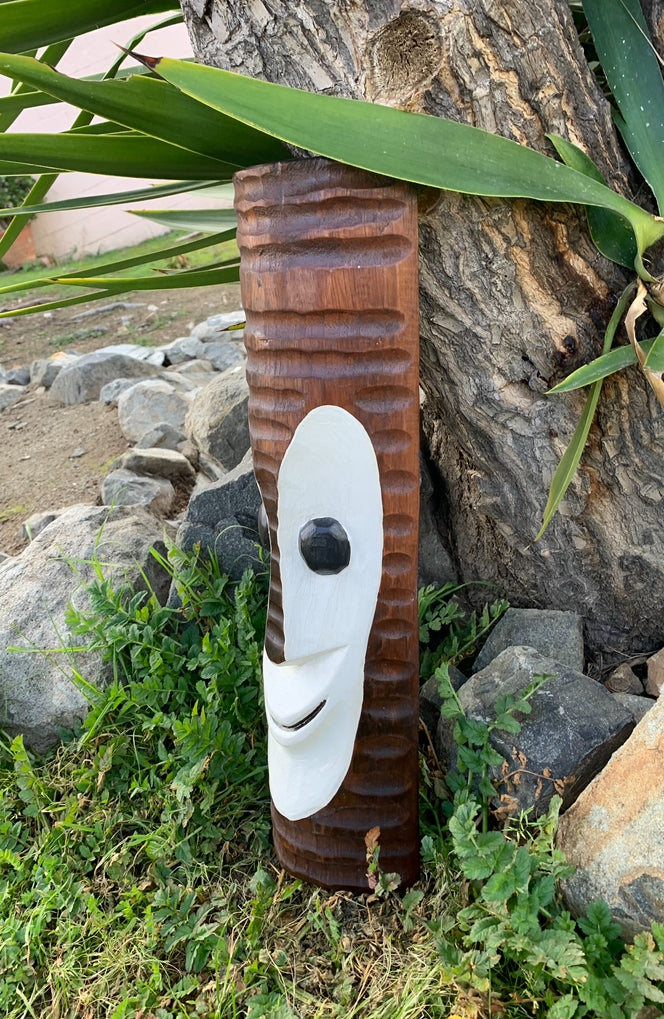 Wooden totem sculpture with a white mask attached, outdoors in a garden beside a tree