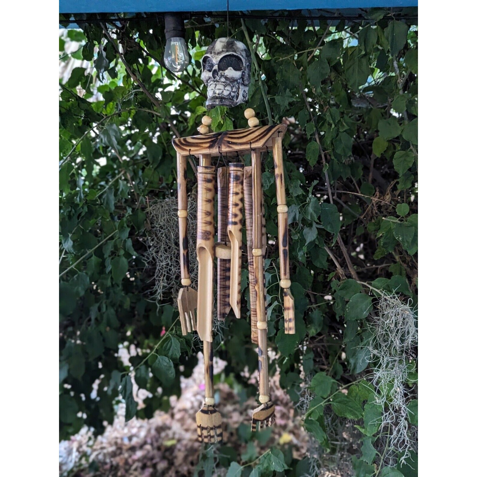 Wooden wind chimes with bamboo tubes and a skull ornament hanging among green leaves.